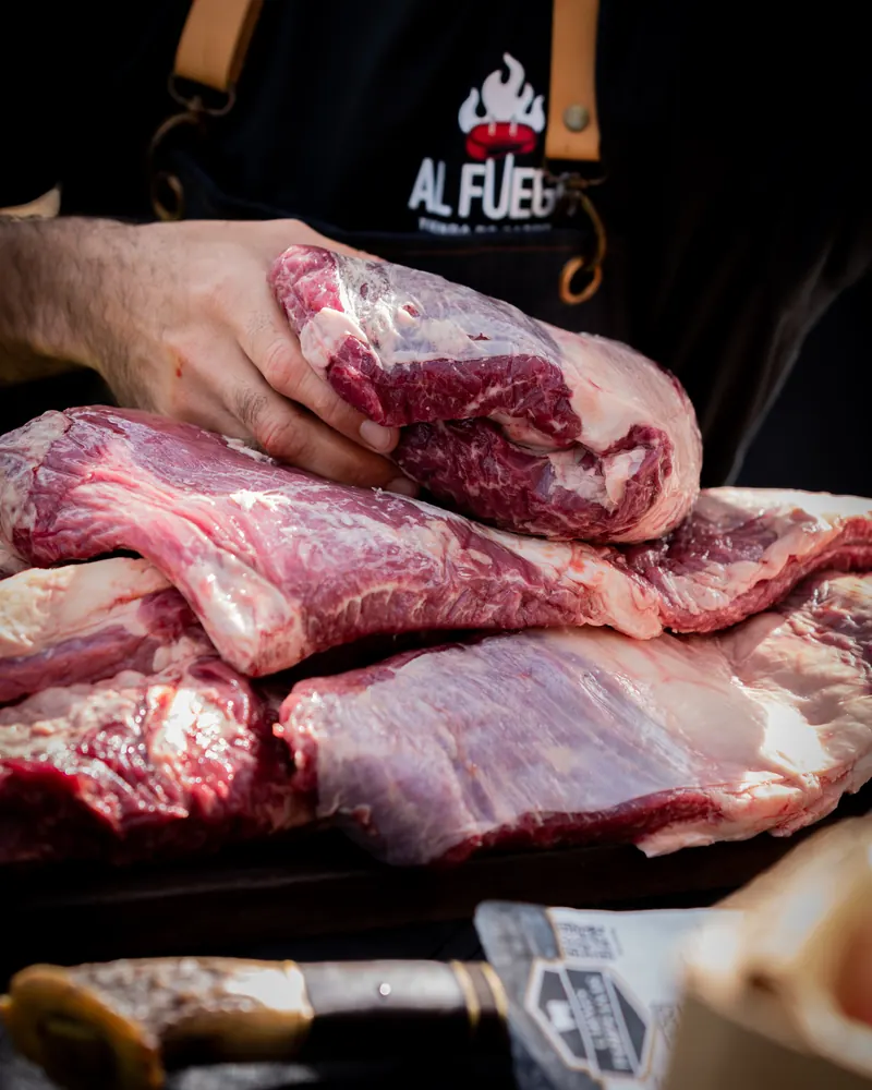 Unas manos, con el delantal de Al Fuego, sujetan piezas de carne cruda sobre una tabla