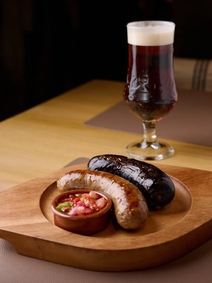 Un chorizo y una morcilla a la brasa sobre una tabla de madera, con un cuenco de salsa y una cerveza