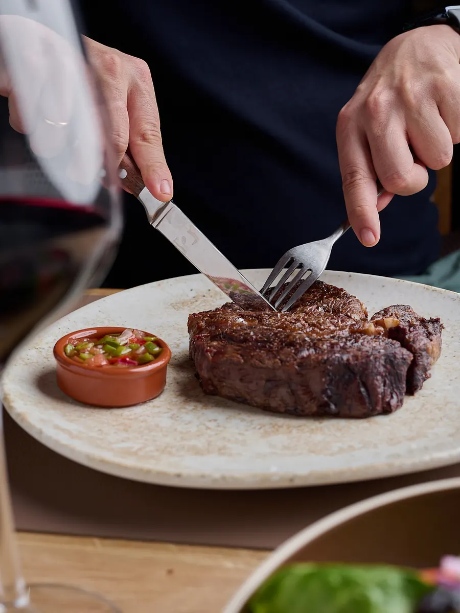 Unas manos cortan un chuletón de carne argentina a la brasa en un plato de cerámica, junto a un cuenco de salsa y una copa de vino tinto