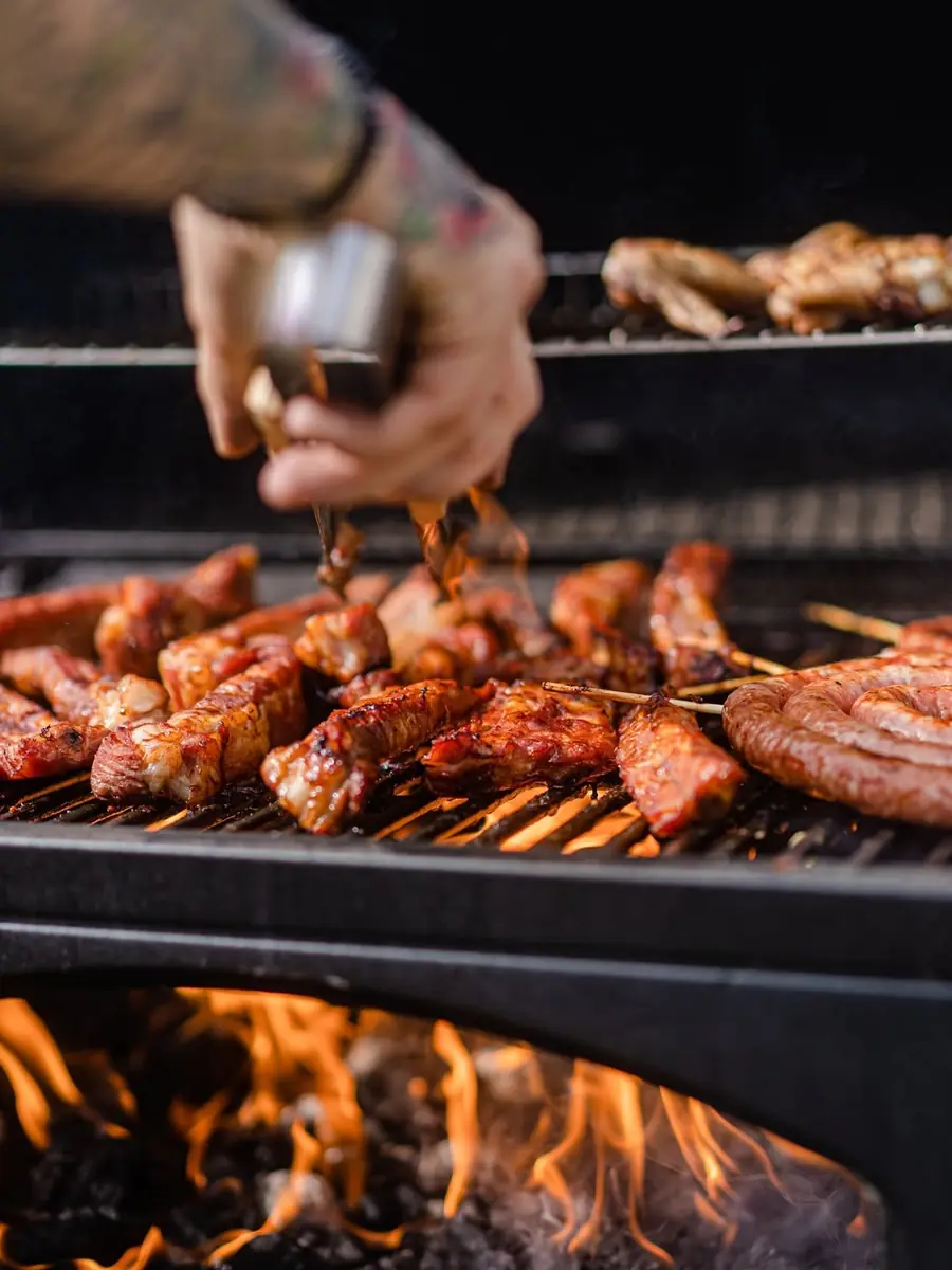 Las manos del parrillero con unas pinzas sobre la parrilla: chorizo, panceta y pollo, con el fuego debajo